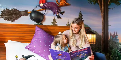 Child and woman sitting on bed reading Room on the Broom in a Room on the Broom Hotel room at the Chessington Resort Hotels celebrating 20 years of Room on the Broom